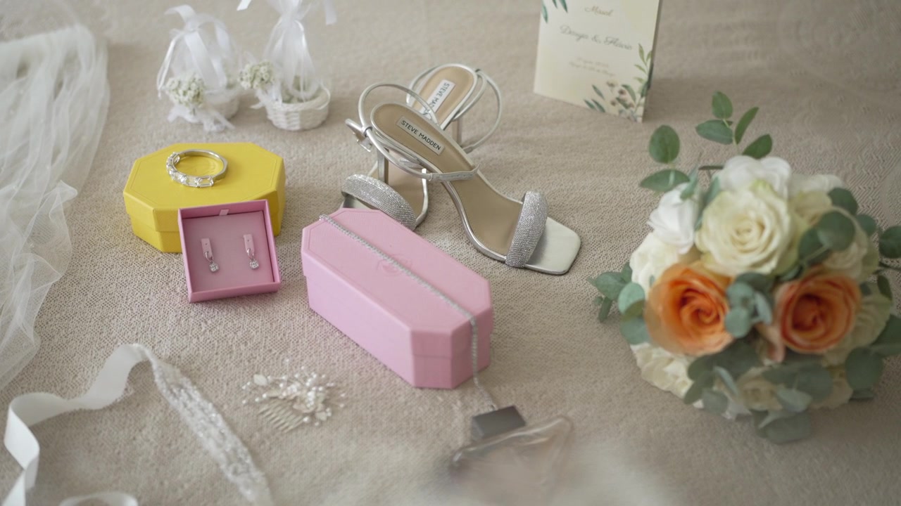 collection of bridal accessories featuring heels, a bouquet with white and orange roses, jewelry boxes, and delicate lace details arranged on a soft beige surface