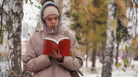 light skin lady dressed in warm winter jacket and beanie standing near tree, reading red book and flipping page while surrounded by autumn foliage and snow-covered ground in peaceful outdoor setting