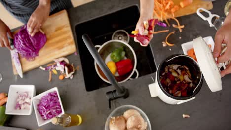 Overhead-of-diverse-couple-in-aprons-preparing-vegetables-composting-waste-in-kitchen,-slow-motion