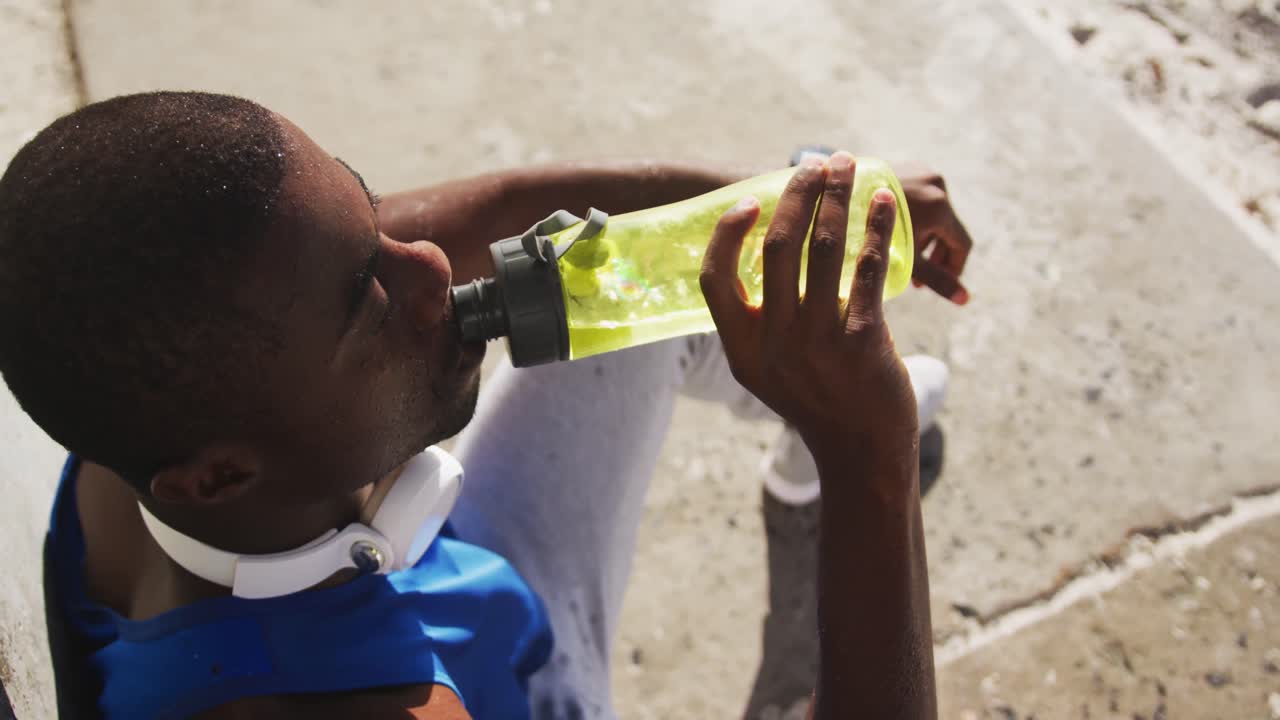 hombre afroamericano sentado y bebiendo de una botella de agua tomando un descanso en el ejercicio al aire libre