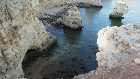 Tilting-up-shot,-Scenic-view-of-Rock-Tunnels-beach-in-Algarve,-Portugal,-people-walking-on-the-coastline-in-the-background