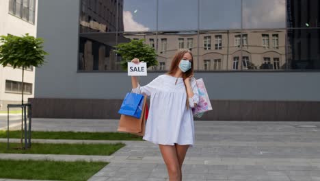 girl in protective mask with shopping bags showing sale word inscription during covid-19 pandemic