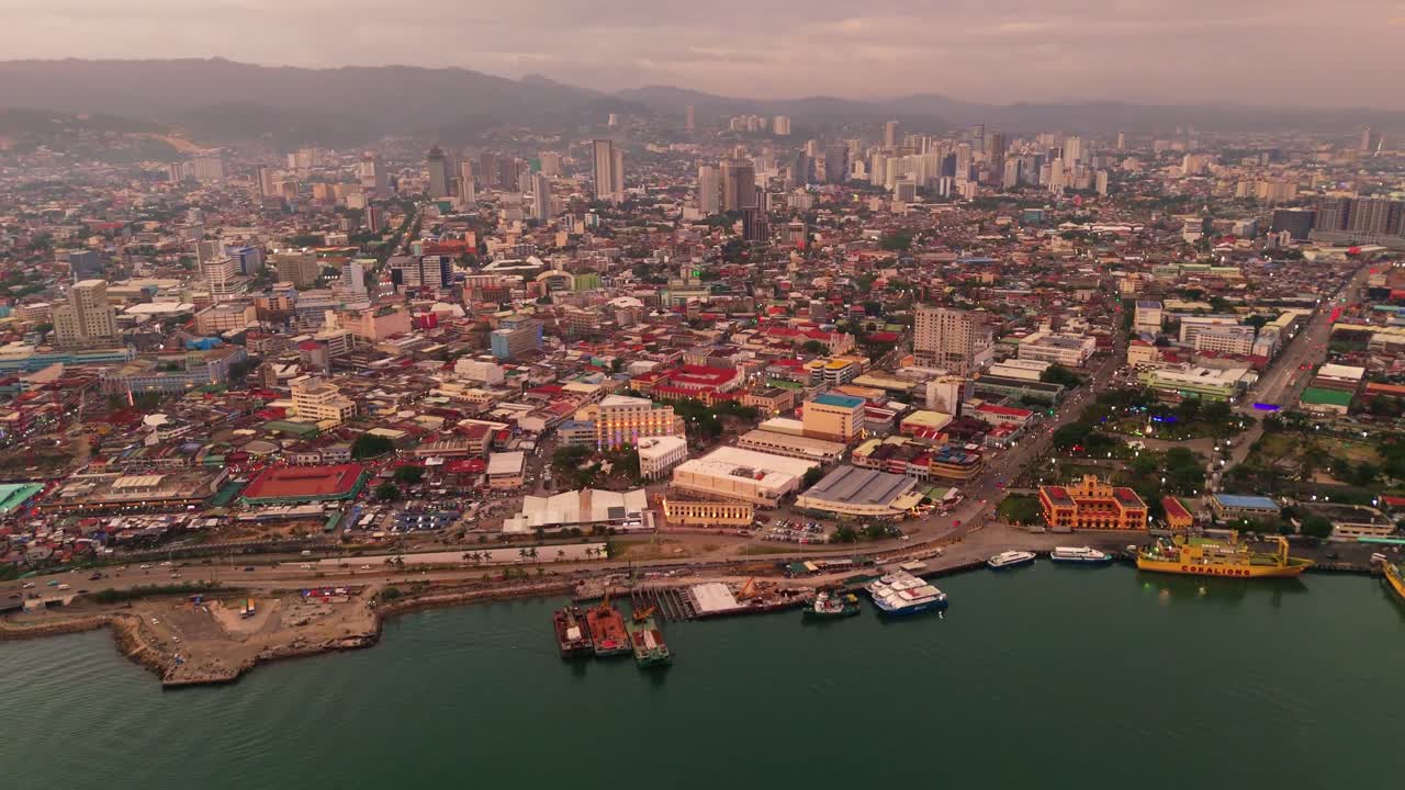 Cebu City, Filippine, panorama aereo al tramonto del paesaggio urbano costiero, caratterizzato dalla trafficata zona portuale, da un denso skyline urbano e da montagne distanti sotto un cielo dorato.