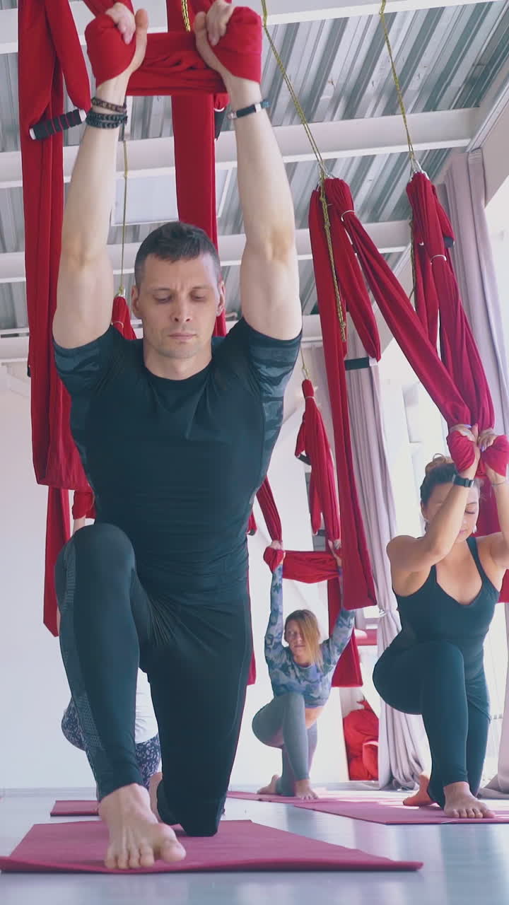 young slim women and man stretch legs holding red aerial fly yoga hammocks above head in modern pilates room