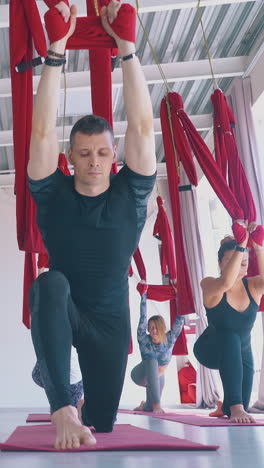 young slim women and man stretch legs holding red aerial fly yoga hammocks above head in modern pilates room