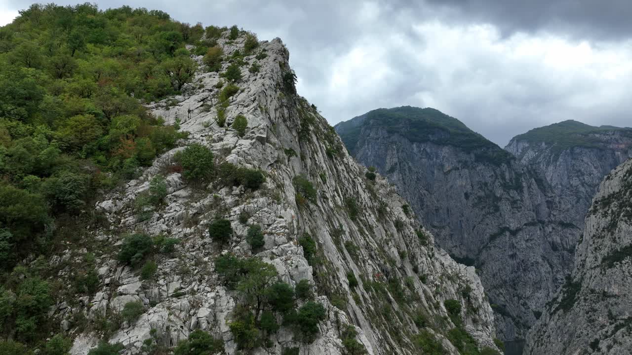 Aerial ascend along mountain pass over Lake Koman, Albania, lush green peaks and deep valleys with white rocks and vegetation spots