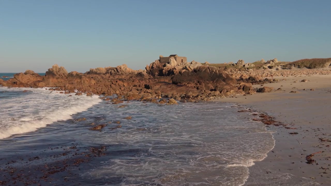 Fremadgående lav droneoptagelse af blide bølger, der skyller ind på en sandstrand med klippekystlinje bagved og krystalklart vand i den sene eftermiddagssol med lyserød granit og militærfort i baggrunden.