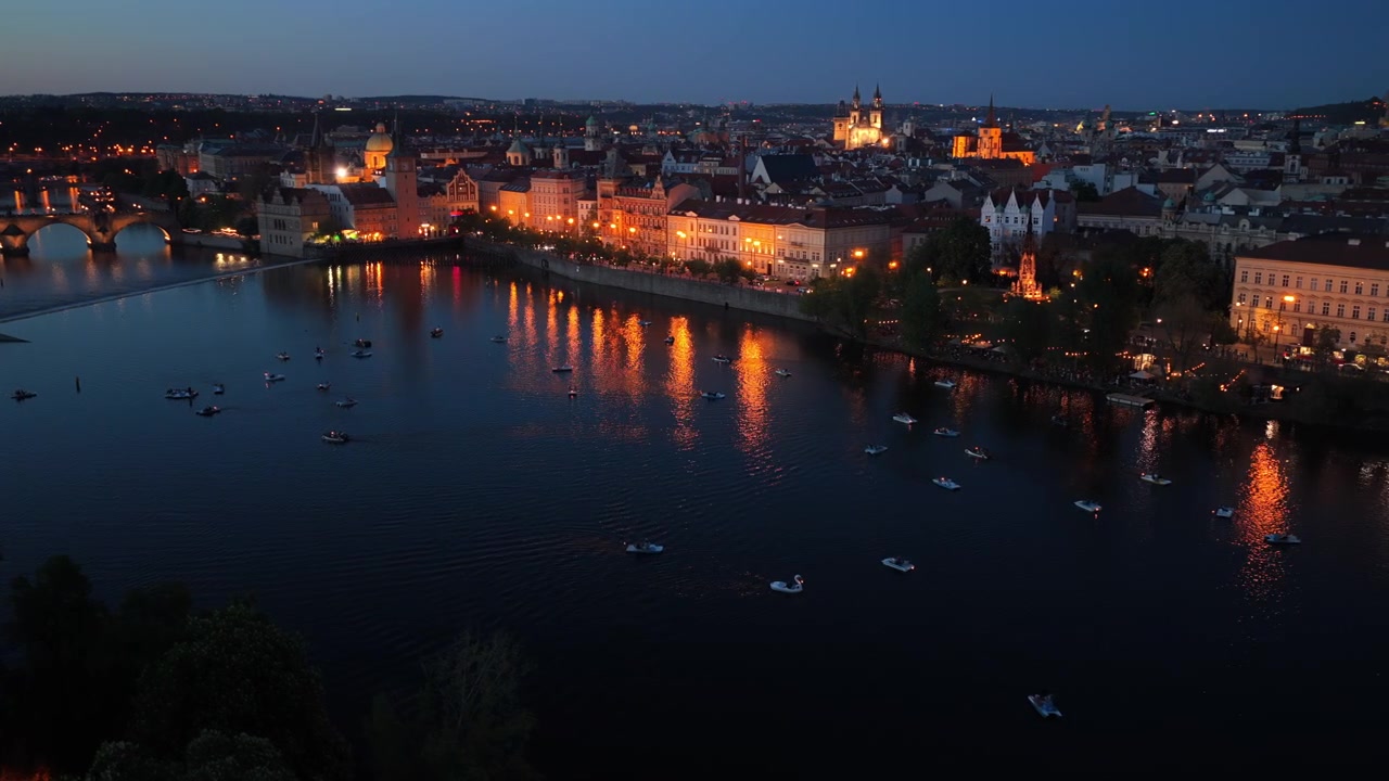 Vista aerea con drone del fiume Moldava di notte con piccole imbarcazioni, il lungofiume illuminato del centro storico e l'arco del Ponte Carlo visibile a sinistra.