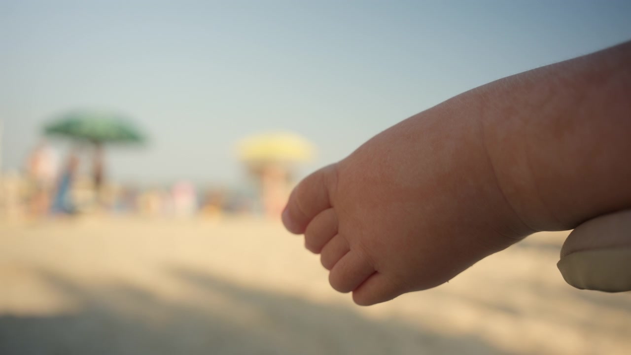 Primo piano di un piedino di neonato a riposo, che si gode una vacanza estiva con sfondo sfocato di ombrelloni da spiaggia e oceano.