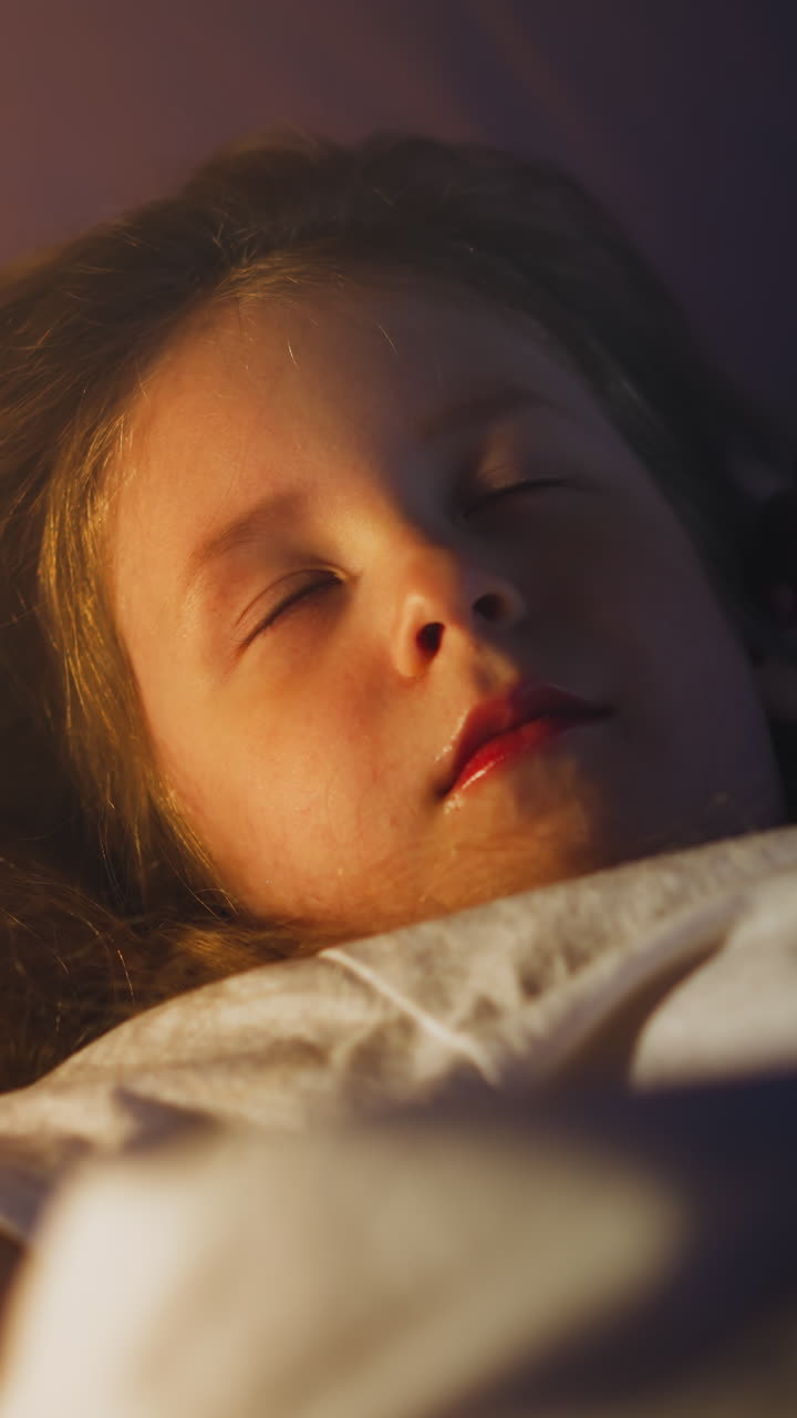 Sweet little girl sleeps with eyes blinking under closed eyelids covered with soft duvet on bed at gentle light of night lamp in dark room slow motion closeup