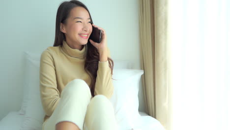 Close-up-pretty-young-woman-sitting-on-a-bed-talking-on-her-smartphone-while-looking-out-the-window