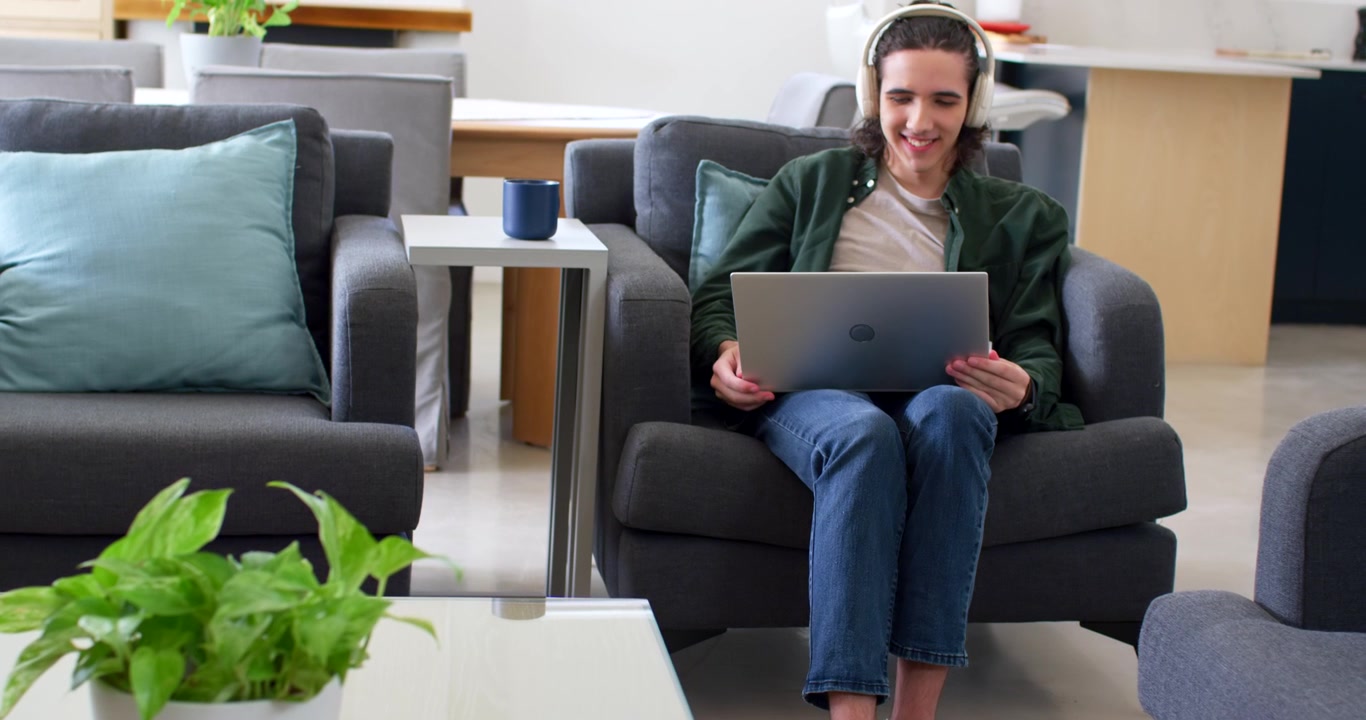 African American man sitting at home in gray armchair wearing white headphones, working on laptop