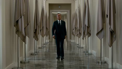 a man in a suit walks down a grand hallway lined with covered flags