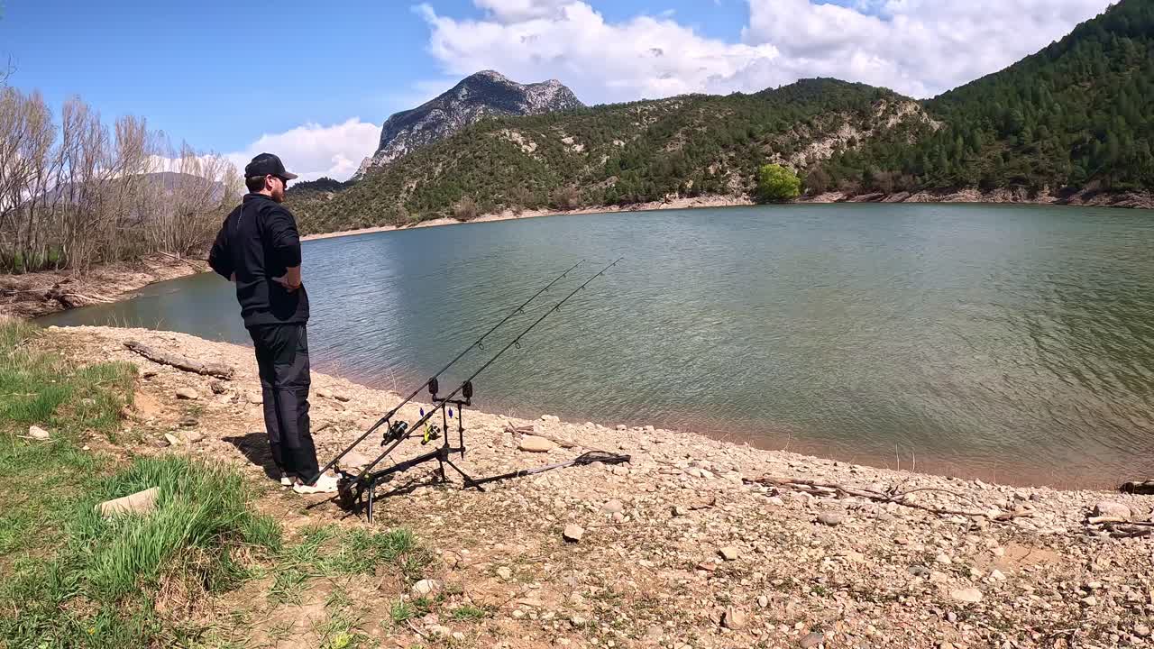 A fisherman stands patiently next to his fishing rods by a tranquil lake, anticipating a bite. The scenic backdrop features lush green hills and a partly cloudy sky