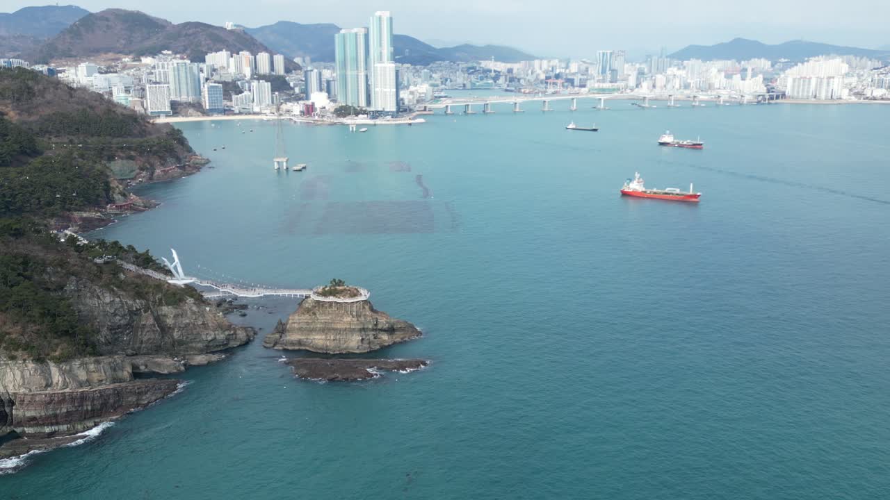 Drone aerial view in South Korea countryside flying over the clear blue sea of Busan next to a green mountain, ships and city in the background sunny day
