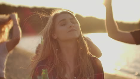 a young girl is dancing on the open air party with her friends. her long blonde hair is flying on the wind. she enjoys a summer evening on the river coast with beer.