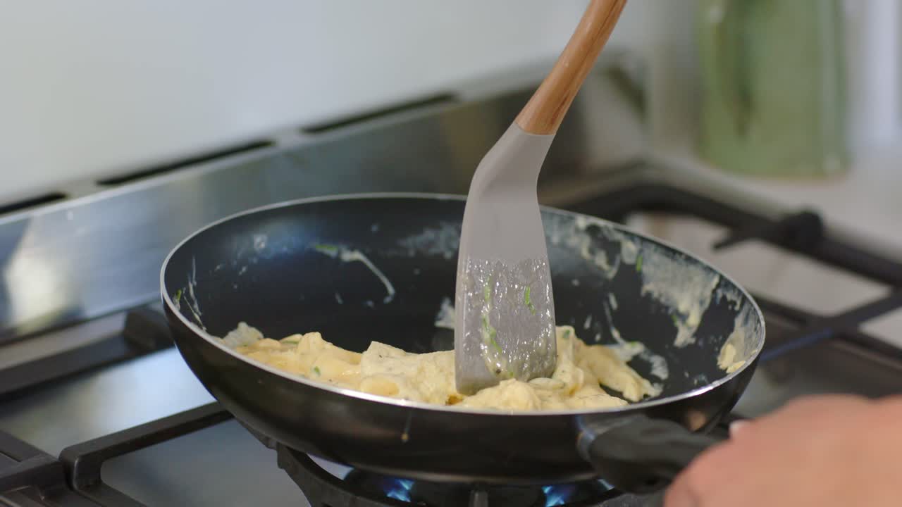 Cook's hand folding eggs on nonstick pan with wooden spatula as gas flame heating for breakfast