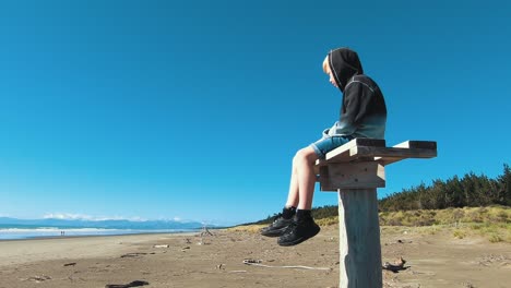 Cámara-Lenta-Alejando-La-Vista-Del-Niño-Sentado-En-Un-Poste-En-La-Playa-De-Madera,-La-Bahía-De-Pegasus,-Mirando-El-Mar-Y-Las-Olas---Tiro-De-Muñeca