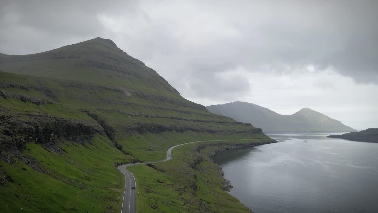 페로 제도의 닝스 (fannings) 에 있는 은 해안 도로, 푸른 언덕과 절벽을 가로질러 휘어지고, 조용한 피오르드를 따라, 구름이 가득한 하늘 아래 극적인 봉우리들로 둘러싸여 평온한 분위기를 만니다.