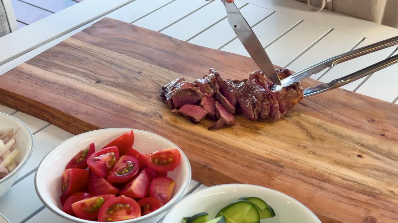 A person uses a chef’s knife and tongs to slice cooked Wagyu beef steak on a wooden cutting board, surrounded by fresh salad ingredients in bright natural daylight