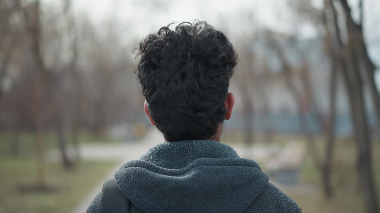 Closeup of young man's head from behind with curly hair and hooded jacket in quiet park during winter, standing still with soft blurred background of trees