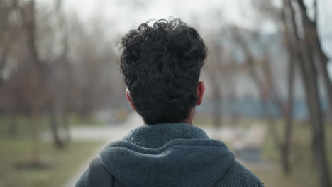 closeup of young man's head from behind with curly hair and hooded jacket in quiet park during winter, standing still with soft blurred background of trees