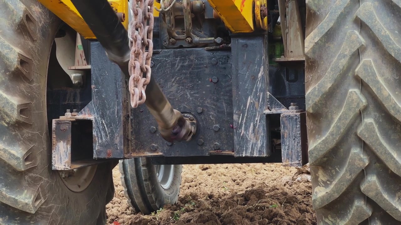 Close view of tractor PTO shaft rotating to power auger drill during ground drilling, mechanical linkage and wheels visible in a rural agricultural setting