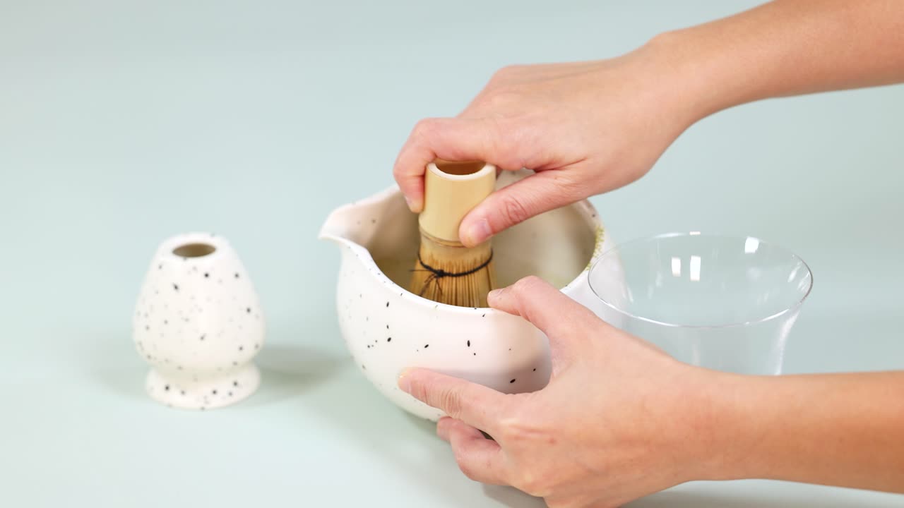 Vigorously whisking matcha powder with bamboo whisk in ceramic bowl under bright studio lighting