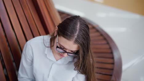 top-down view of young adult wearing glasses, head slightly bent, deeply focused on something while seated on wooden bench, soft lighting and wooden texture create a calm, introspective ambiance