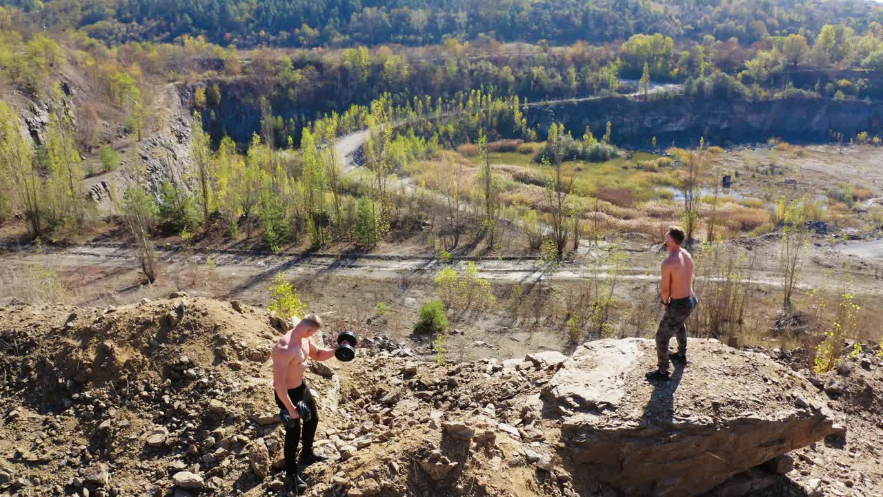 Sportsmen training with sports equipment among nature. Two muscular athletes doing exercises with dumbbells on the top of a hill. Aerial view.