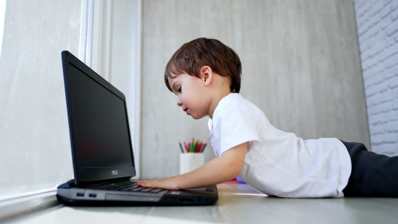 Small kid lies down on the floor in front of laptop. Toddler dislikes his pose and rises closing the gadget. Low angle view.