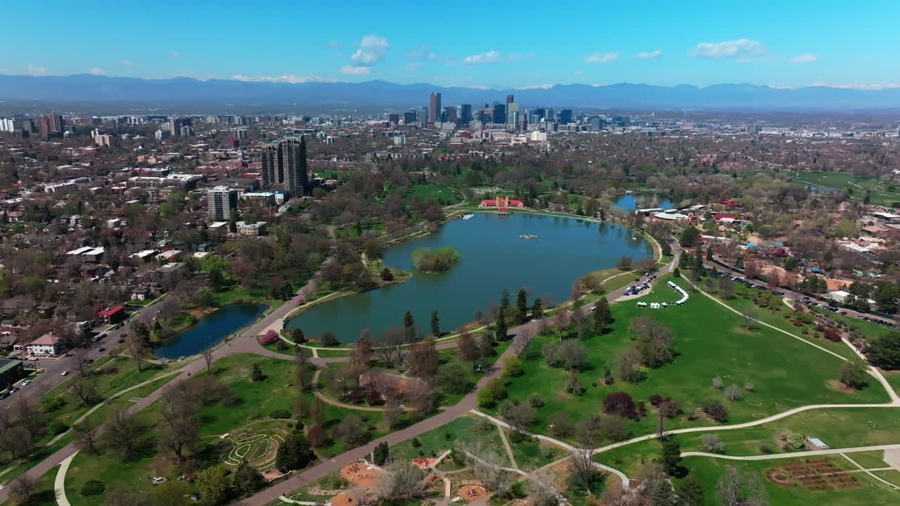 Leo City Park Denver Colorado Lake Pavilion vibrant spring summer aerial drone front range cityscape green lush grass trees blossom sunny blue sky snow capped Rocky Mountains circle left motion