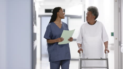 African-american-female-doctor-and-senior-woman-with-crunches-walking-in-hospital,-slow-motion