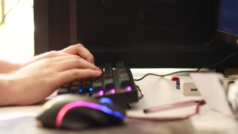 toma lateral de las manos de un hombre interactuando con un teclado rgb en un escritorio de computadora sucio