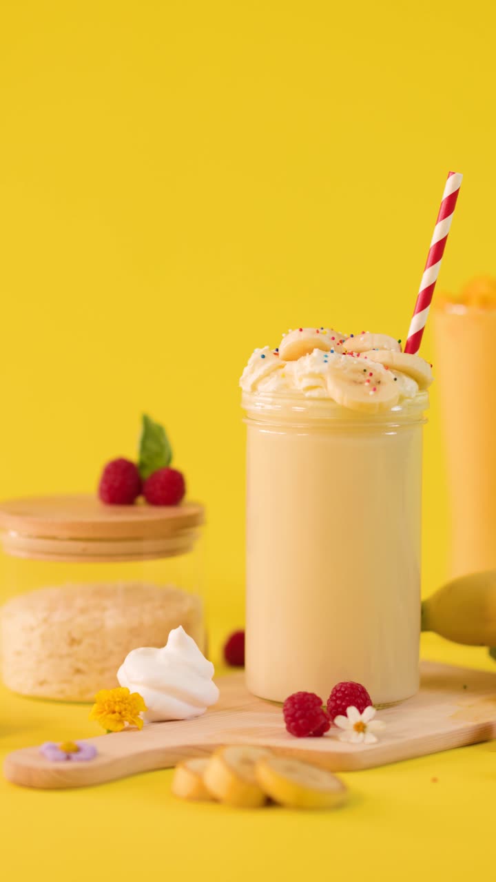 Bicchiere di frullato alla banana con panna montata, mano che afferra una cannuccia a righe, con illuminazione da studio colorata