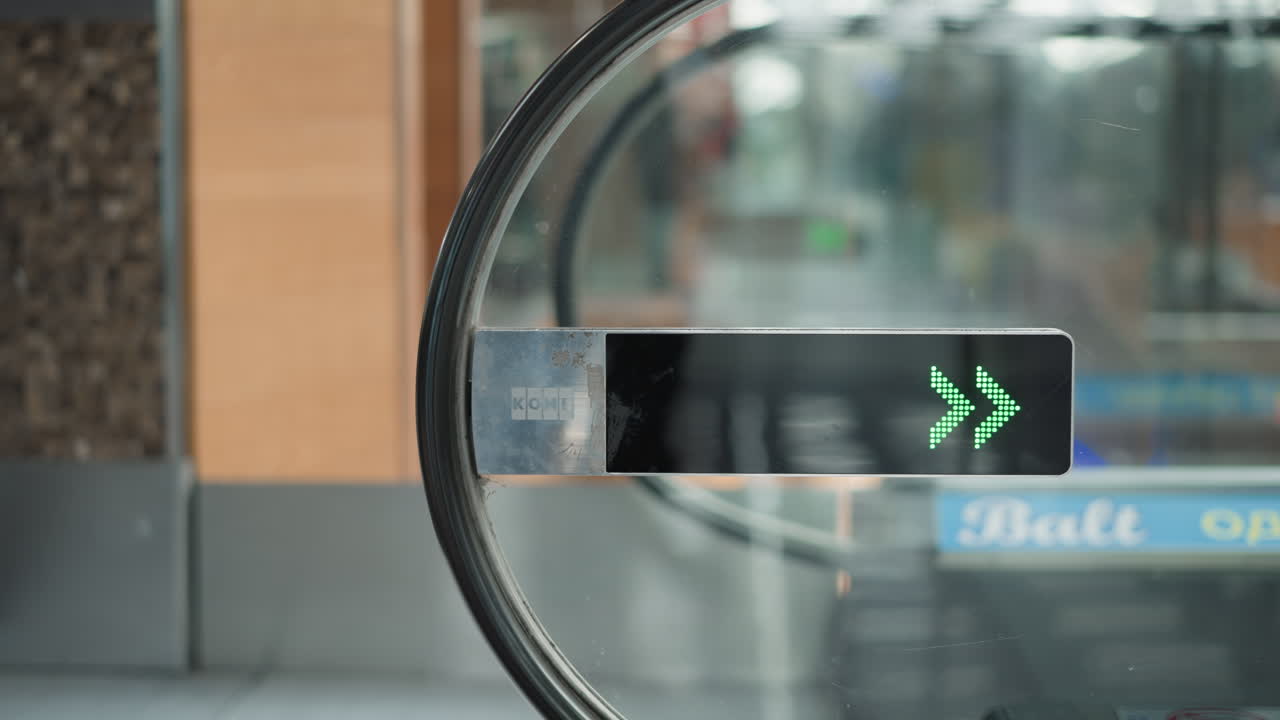 side view of circular glass mounted green led arrow display pointing down at moving walkway entrance with safety icons on side panel and blurred background of bright mall interior