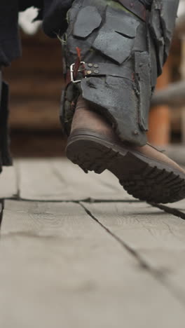 man in heavy boots with buckles walks slowly along wooden platform past fence. warrior in metal armor patrols medieval settlement closeup on blurred background