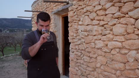 Ein-Lächelnder-Bärtiger-Mann-Stößt-Mit-Einem-Glas-Weißwein-Im-Hintergrund-Einer-Typisch-Mediterranen-Steinhütte-An