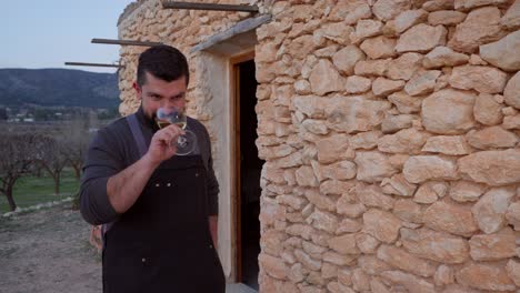 un hombre barbudo sonriente brinda con una copa de vino blanco en el fondo de una típica cabaña de piedra mediterránea