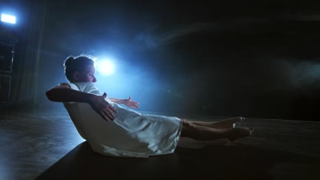 modern dance woman in a white dress dances a modern ballet, jumps, makes rotation on the stage with smoke in the blue spotlights.