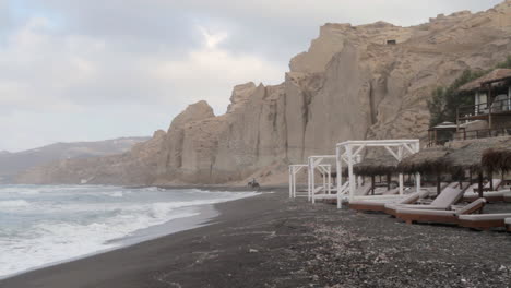 Ein-Reiter-Reitet-Auf-Seinem-Pferd-Am-Schwarzen-Strand-Von-Santorini-Mit-Weißen-Vulkanischen-Klippenformationen-Im-Hintergrund