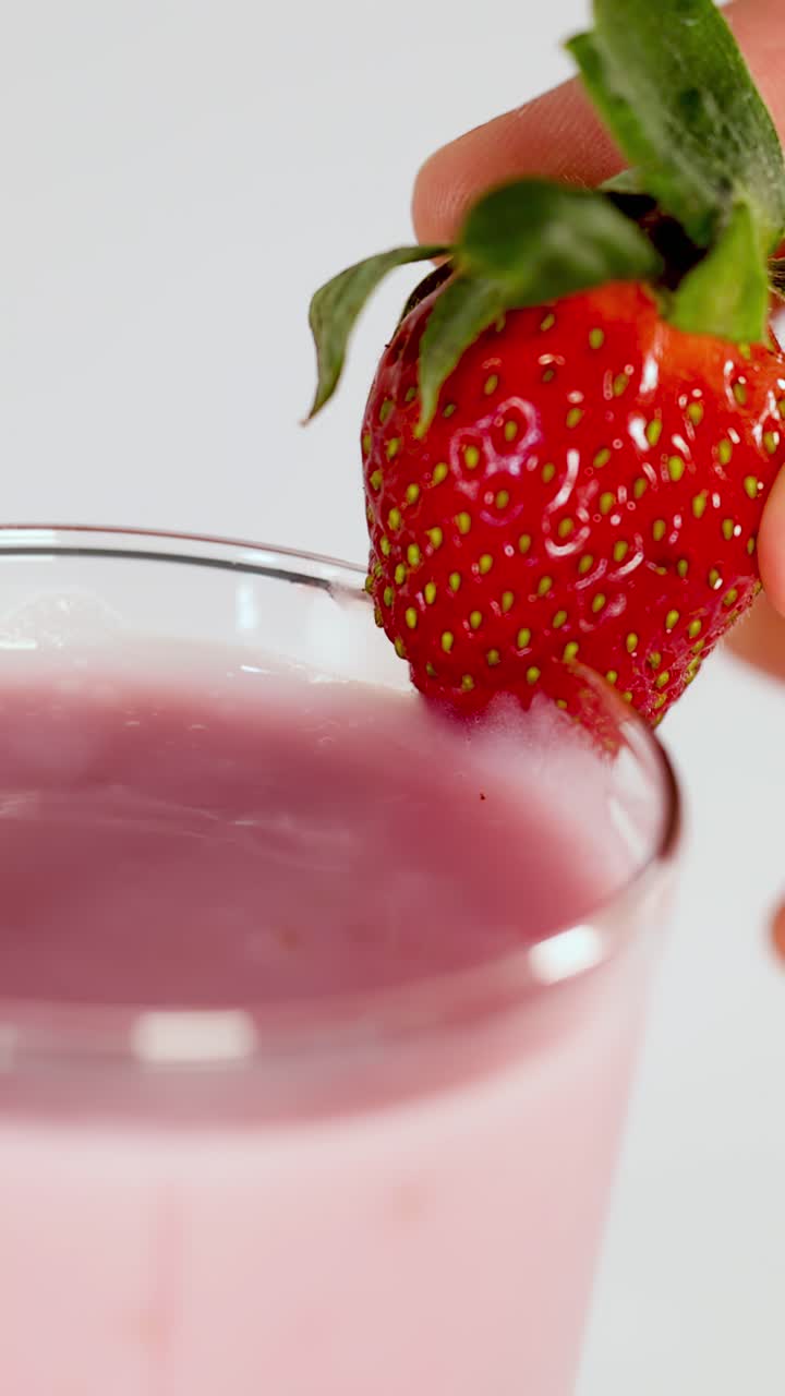 Primo piano di una mano che posiziona una guarnizione di fragola fresca su un frullato di fragole rosa su uno sfondo pulito.