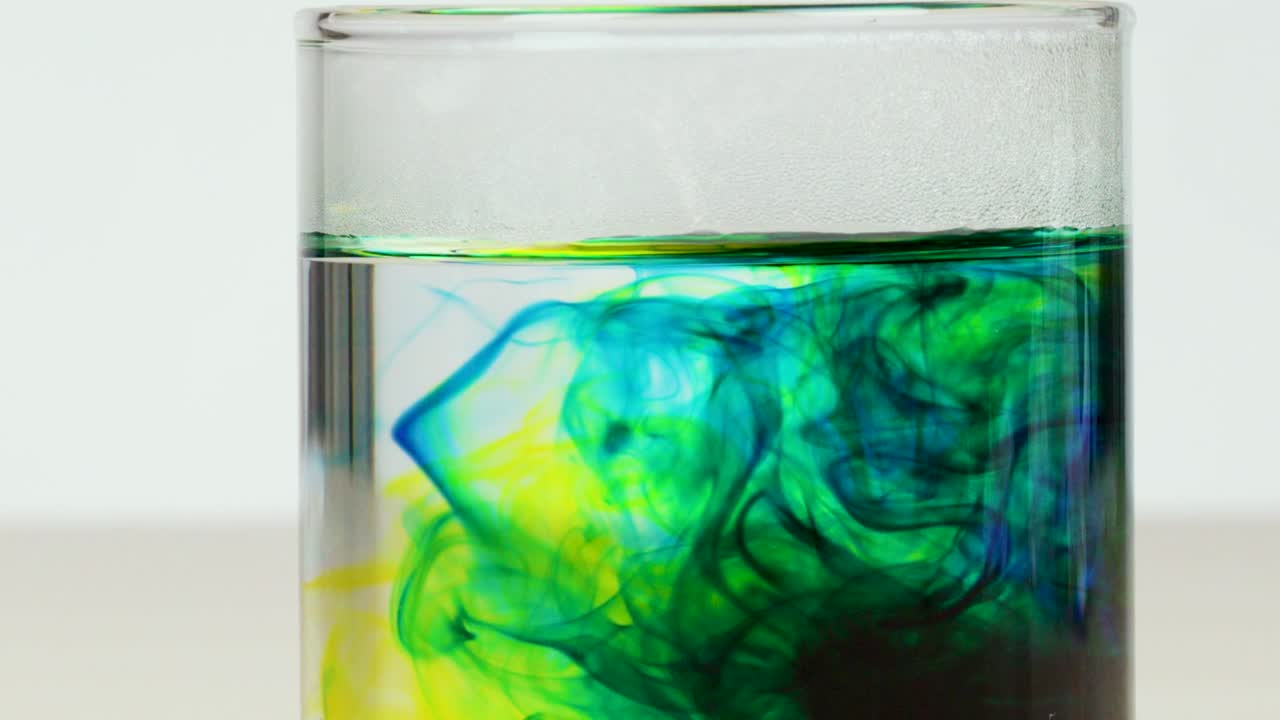 Vibrant blue and yellow food coloring diffuses and mixes inside a clear glass of water