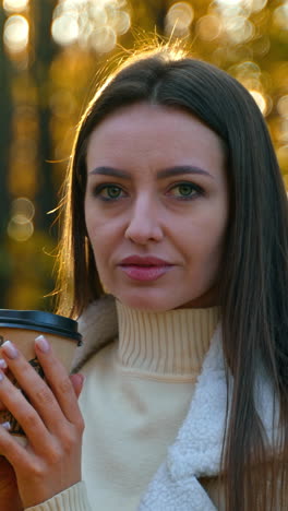 dark-haired lady drinking tea from paper cup outdoors. lady looks at camera, touching her hair and looking for a moment away. close up. blurred autumn backdrop.