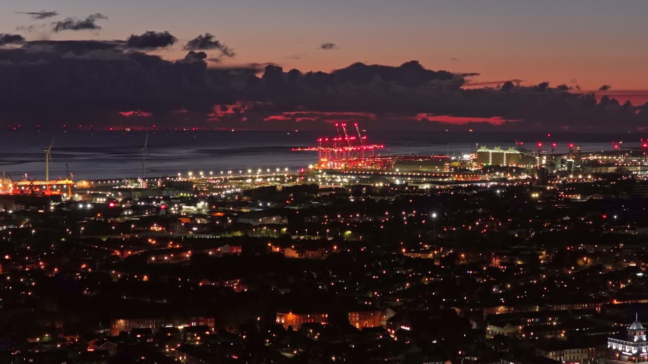 Drone static over Liverpool’s waterfront at night, revealing glowing cranes, wind turbines, and city streets—showcasing the city’s mix of industry, energy, and urban life under twilight skies