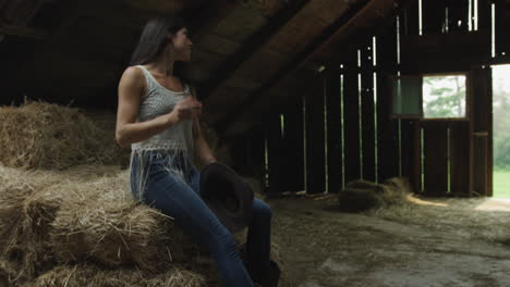 confident young cowgirl in a barn puts on her cowboy hat and gloves, picks up a bale of straw and carries it