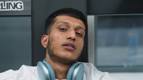 young man sitting against gym bench with tired expression resting after intense workout session wearing white sportswear and wireless headphones around neck in bright modern fitness environment