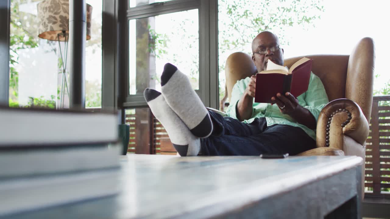 Happy african american senior man relaxing in armchair with feet up, reading a book