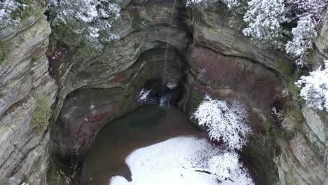 Rising-drone-of-waterfall-in-beautiful-cliff-formation-rising-reveal-vast-winter-wonderland,-snow-covered-tree-tops