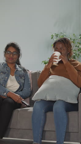 indian and african american women watch movie together sitting on sofa in living room. female friends talk holding remote control and cup of tea